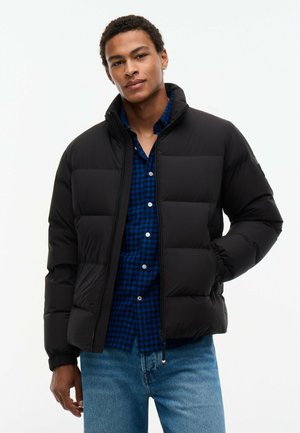 Joven con una chaqueta acolchada negra sobre una camisa a cuadros azul y vaqueros azul claro, de pie con una mano en el bolsillo.