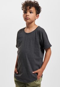 Dreng med krøllet hår iført en mørkegrå t-shirt med oprollende ærmer og lysegrønne bukser, stående med hænderne i lommerne foran en ensfarvet baggrund.