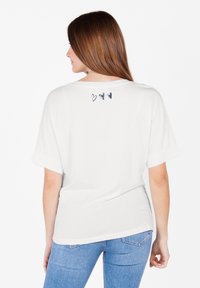 Camiseta blanca de manga corta con escote redondo y tres pequeños corazones oscuros impresos en la parte superior de la espalda, combinada con jeans azules.