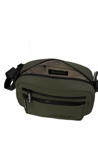 Bolso cruzado en verde oliva con múltiples bolsillos con cremallera, exterior liso y acentos de hardware negro. Detalle del logo reflectante en la parte frontal.