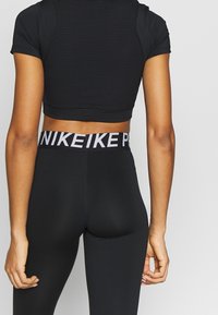 Musta lyhytcroppinen urheilu-toppi, jossa on ribbikuvio ja lyhyet hihat, yhdistettynä mustiin leggingseihin, joissa on valkoinen "NIKE"-logovö ja.