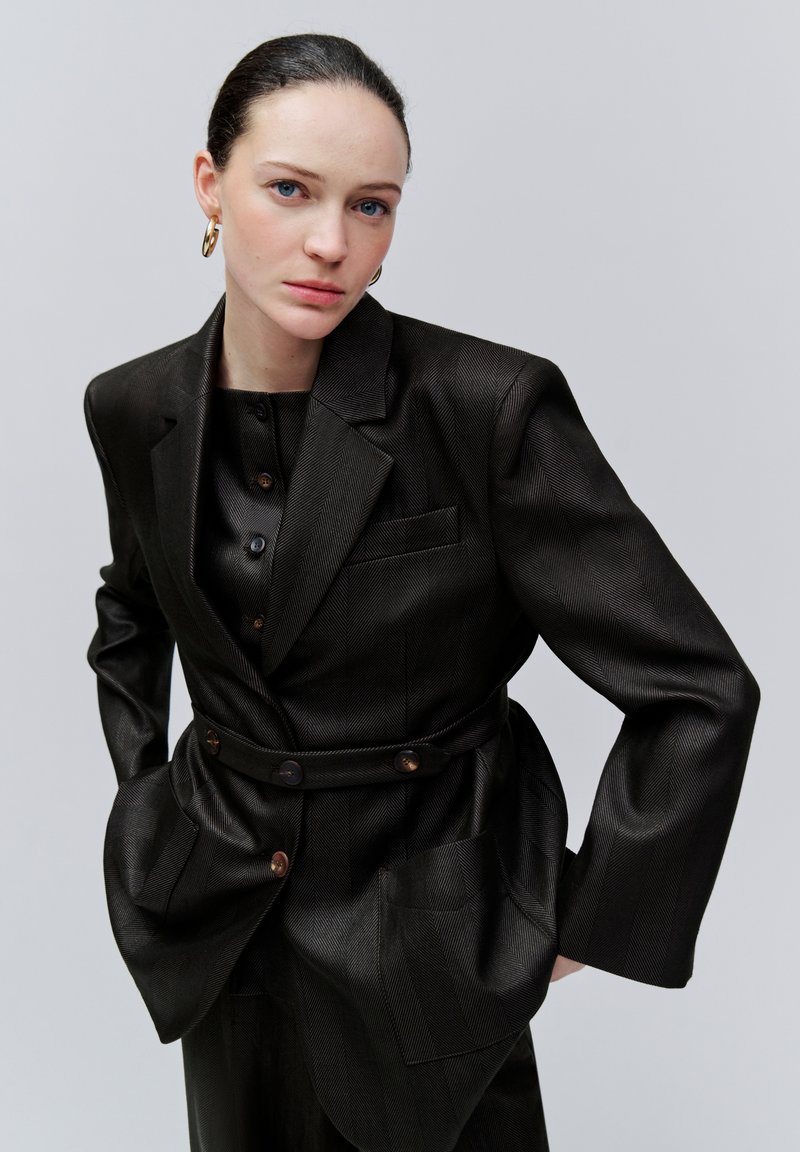 Femme à la peau pâle et aux yeux bleus, portant un blazer noir texturé et une chemise, les mains dans les poches, sur un fond gris clair uni.