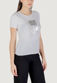 Camiseta blanca de manga corta con un diseño de logotipo plateado brillante en la parte frontal. La tela parece suave con un corte ajustado. Combinada con pantalones negros.