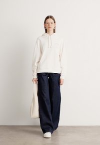 Felpa bianca con cappuccio e cordini, dotata di tasca frontale, abbinata a jeans in denim scuro a gamba larga. La modella porta una borsa tote beige e indossa scarpe bianche.