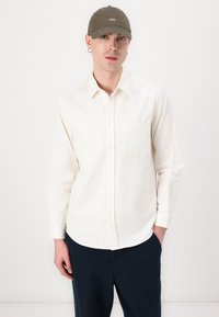 White textured button-up shirt with a chest pocket, worn with dark pants, complemented by an olive cap featuring white stitched lettering.