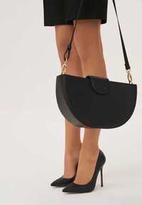 Fekete bőr félhold alakú kézitáska arany színű hardverrel és pántos záródással. Magas sarkú fekete cipők háttere előtt elhelyezve.