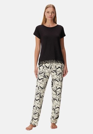 Vrouw staat blootsvoets, draagt een zwart kort mouwen topje met kanten rand en witte broek met zwarte abstracte bladpatronen.