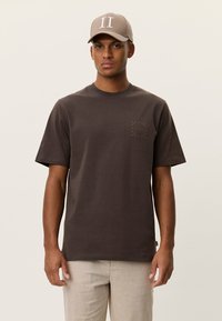 Καφέ βαμβακερό μπλουζάκι με κοντά μανίκια, λαιμόκοψη τύπου crew και μικρή γραφιστική λεπτομέρεια στο στήθος. Φορεμένο με μπεζ καπέλο που φέρει λογότυπο.