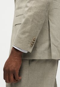 Σκούροχρωμο χέρι που ακουμπά δίπλα στο μανίκι ενός μπεζ σακακιού ψαροκόκαλο με τέσσερα κουμπιά και αντίστοιχο παντελόνι.