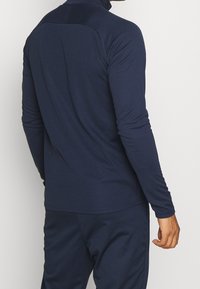 Sportowy top z długim rękawem w kolorze ciemnego granatu, z wysokim kołnierzem i dopasowanym krojem, o gładkiej fakturze i minimalną ilością szwów na plecach.