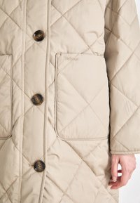 Manteau matelassé beige avec de gros boutons en écaille, deux poches avant et le logo "Barbour" visible sur la poche droite, porté par une personne.