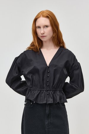 Femme aux longs cheveux roux portant un chemisier en satin noir avec des boutons et des manches bouffantes, associée à une jupe en jean noire, posant devant un fond uni.