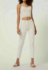 Vrouw in een mouwloze crème crop top met kant, hoge witte jeans en nude geweven open teen blokhak sandalen.