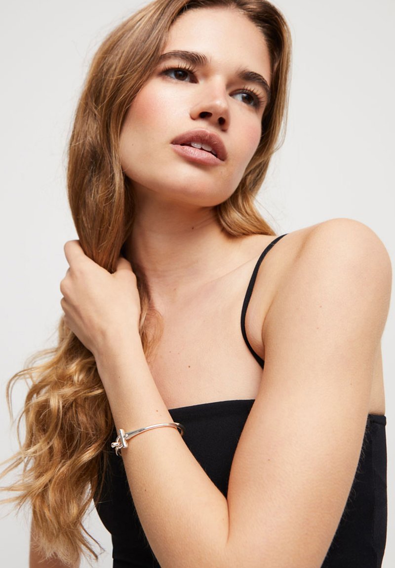 Bracelet en métal argenté avec un accent de nœud délicat, porté sur un bras bronzé. L'arrière-plan présente une couleur neutre, mettant en valeur le design du bracelet.