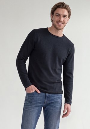 Mann trägt ein schwarzes Langarmshirt und blaue Jeans, eine Hand in der Hose, lächelt vor einem schlichten hellen Hintergrund.