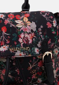 Blommig rygsäck i svart tyg, med en guldlogotyp och svarta läderremmar. Designen inkluderar färgglada blommor och betydande detaljer.