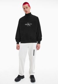 Felpa nera con collo alto con grafica "Calvin Klein Jeans"; abbinata a pantaloni cargo bianchi con tasca con zip e scarpe nere con lacci.