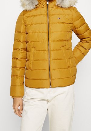 Mujer que lleva una chaqueta acolchada amarilla mostaza con cremallera frontal y capucha con borde de piel, combinada con pantalones de color claro.