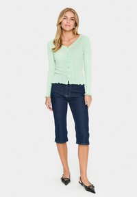 Cardigan côtelé vert clair avec un col en V et une fermeture par boutons, associé à un jean bleu foncé jusqu'aux genoux et des ballerines noires à bout pointu.