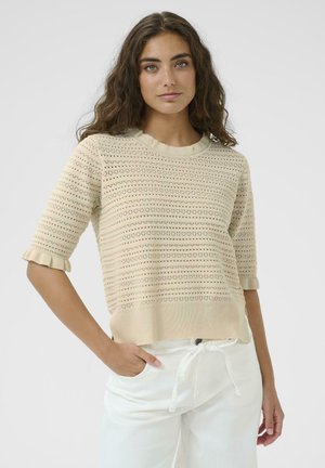 Beige knitted sweater with short, ruffled sleeves, featuring a textured pattern of small holes and horizontal stripes. Worn with white pants.