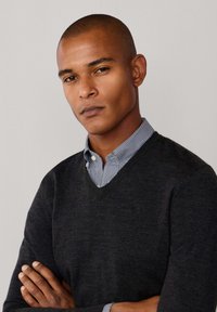 Donkergrijze v-hals trui gedragen over een lichtblauw en wit geruite overhemden met kraag. Gladde textuur, aansluitend ontwerp. Geen tekst of logo's zichtbaar.