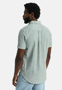 Camicia a maniche corte a righe verdi e bianche, realizzata in tessuto leggero. Presenta un'apertura frontale e un orlo arrotondato sul retro.