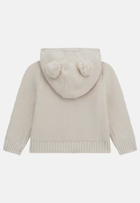 Haut en tricot couleur crème avec des poignets et un ourlet côtelés. Présente deux pompons à l'arrière de la capuche. Motif texturé sur l'ensemble.