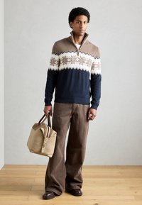 Camisola de malha azul-marinho e creme com padrão de flocos de neve, detalhes em bege, combinada com calças largas castanho e uma mala castanha com alças castanhas.