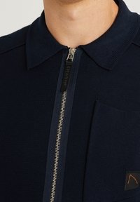 Marinblå zip-up skjorta i texturerad tyg, med krage, bröstficka och en kontrasterande dragkedja med en svart etikett.