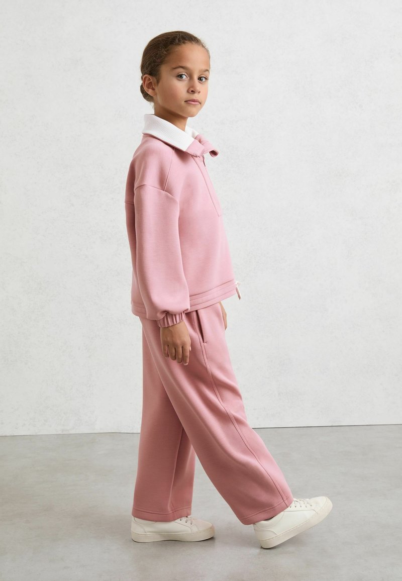 Jeune fille marchant, portant un sweat-shirt rose avec col blanc, un pantalon rose ample et des baskets blanches sur un fond uni.