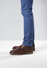 Braune Leder-Stiefeletten mit strukturierter Oberfläche, elastischen Seiteneinsätzen und robusten Gummisohlen, kombiniert mit blauen, eng anliegenden Hosen.