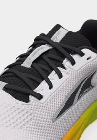 Κοντινό πλάνο ενός λευκού και μαύρου αθλητικού παπουτσιού με μαύρα κορδόνια, επάνω μέρος από διαπνέον πλέγμα και ενδιάμεση σόλα με κίτρινη διαβάθμιση, που φέρει το λογότυπο Altra.