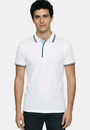 Jeune homme aux cheveux noirs et bouclés portant un polo blanc avec des bordures noires et bleu clair, debout devant un fond uni.