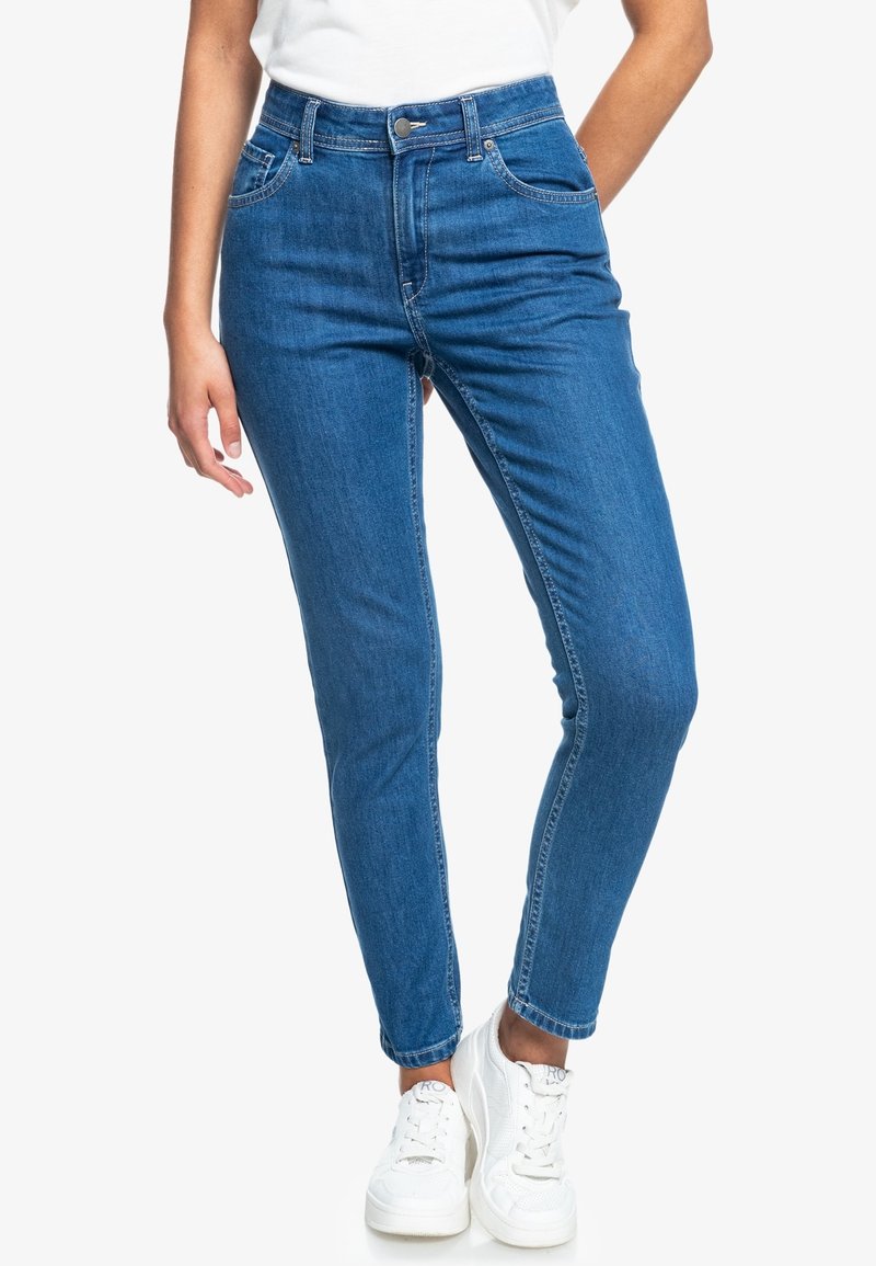 Jean slim bleu en denim taille mi-haute avec fermeture par bouton et zip, porté avec des baskets blanches et un top blanc rentré à l’intérieur.