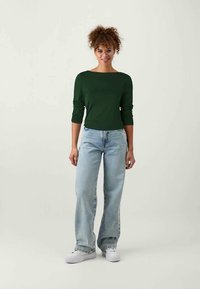 Giovane donna con capelli ricci e occhiali, indossa un maglione verde scuro a maniche lunghe, jeans a gamba ampia azzurri e sneakers bianche, in posizione eretta.