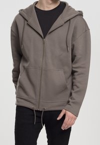 Uomo che indossa una felpa con zip color taupe, con tasche frontali e cordini, abbinata a pantaloni neri su uno sfondo chiaro.