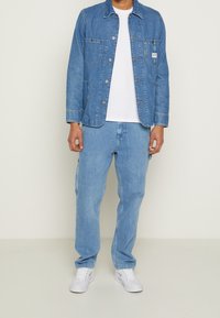 Jeansjacka i ljusblått med två framfickor, knäppning med knappar och en vit t-shirt under. Kombinerad med matchande ljusblå jeans och vita sneakers.