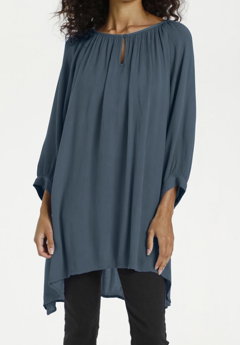 Femme portant une tunique bleue ample à manches longues avec une petite fente à l'encolure avant, associée à un pantalon noir sur fond blanc.