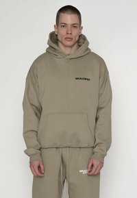 Felpa con cappuccio color khaki con tasca frontale e logo nero "MULTIPLY". Tessuto morbido e strutturato, vestibilità ampia, abbinata a pantaloni della tuta coordinati.