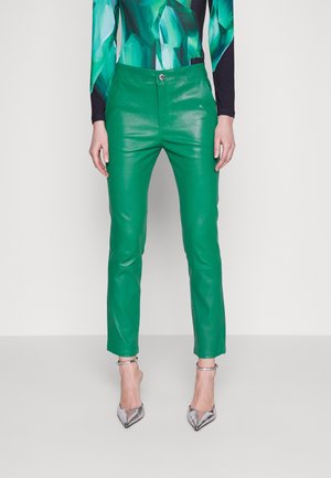 Pantalon en cuir vert avec une coupe ajustée, doté d'une fermeture à bouton à l'avant et d'une texture lisse, associé à des chaussures à talons hauts argentées.