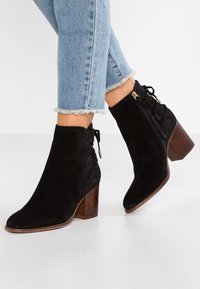 Botas de tobillo de ante negro con punta cuadrada, tacón grueso de madera, cremalleras laterales y detalles de encaje decorativos. Los jeans tienen dobladillos deshilachados.