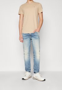 Beige puuvillainen t-paita yhdistetty vaaleansinisiin farkkuihin, jotka esittelevät haalistuneen tekstuurin ja hienovaraisen sävyn, viimeistelty valkoisilla tennareilla.