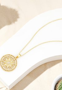 Guldtonad halsband med en cirkulär berlock med intrikat utskärningsdesign och stenprydda accenter, hängande från en delikat kedja.