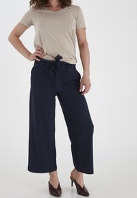 Beige tričko s krátkým rukávem a výstřihem do V, spárované s námořnickými kalhotami s širokými nohavicemi a zavázaným pasem. Modelka má na sobě hnědé sandály na podpatku.