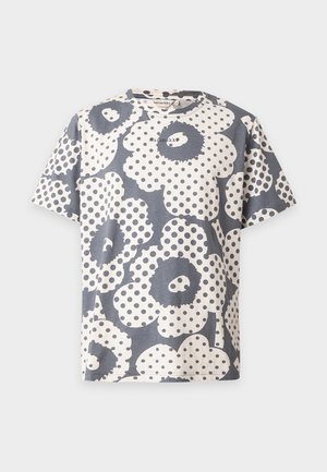 Mann steht mit den Händen in den Taschen, trägt ein passendes schwarz-weißes T-Shirt und Hosen mit Blumenmuster sowie graue Sneakers auf einfarbigem Hintergrund.