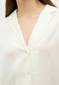 Blouse blanche semi-transparente avec col cranté, fermeture à un bouton et texture lisse. Design simple et classique avec des détails minimaux.