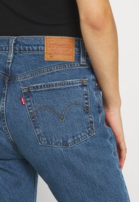 Jeans de mezclilla azul con un ajuste clásico, que cuentan con una etiqueta de cuero marrón en la cintura y costuras arqueadas distintivas en el bolsillo trasero.