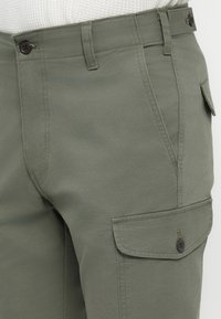 Pantalons cargos verts olive en tissu robuste, avec poches avant, une poche à rabat sur le côté et des boutons foncés pour la fermeture.
