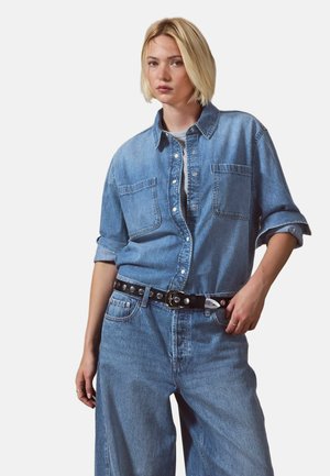 Kvinde med blondt hår iført en lyseblå denimskjorte med opkørte ærmer, vide jeans og et sort bælte med nitter.