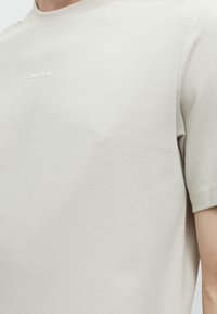 Κοντινό πλάνο μπεζ κοντομάνικου πουκάμισου με μικρό λευκό λογότυπο "Calvin Klein" στο στήθος, που φοράει ένα άτομο.